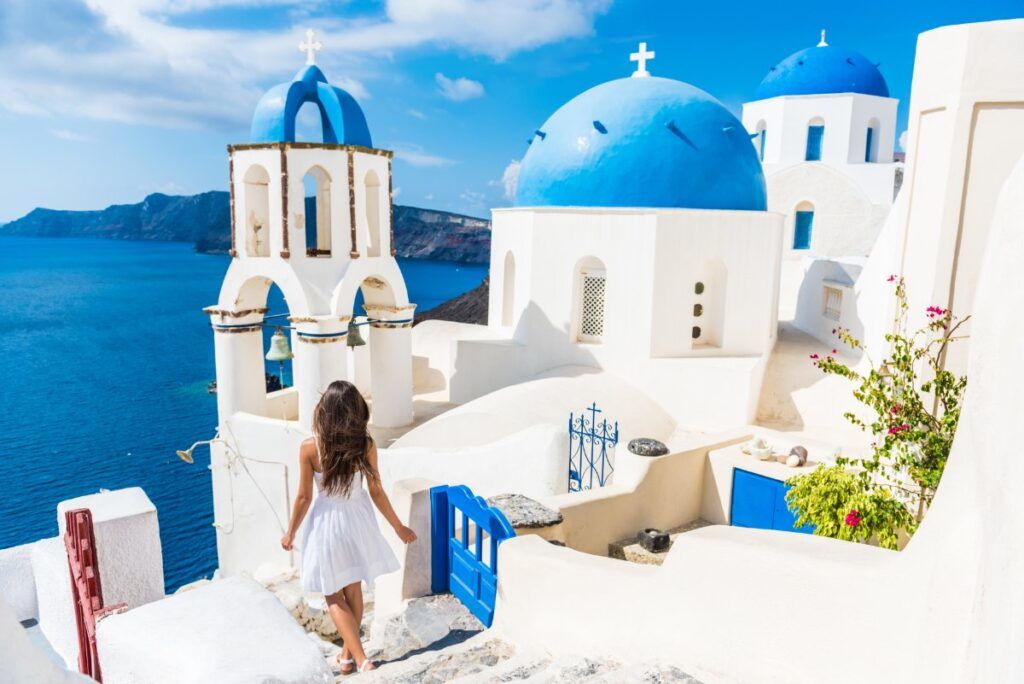 a woman walks through the narrow streets of Santorini between white buildings and blue sea