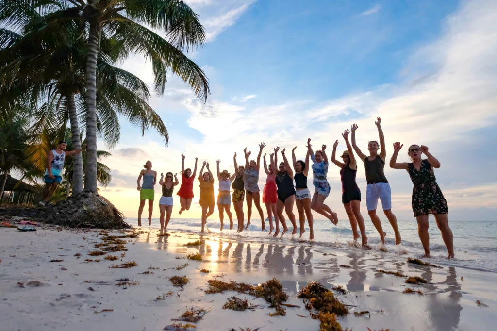 A large group of WeRoad travelers jumping for joy on a white sand beach at sunset in Cuba.