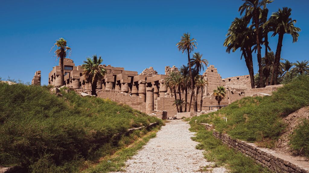 Gravel path leading through grassy mounds towards the imposing ancient ruins and columns of the Karnak Temple complex, flanked by palm trees.