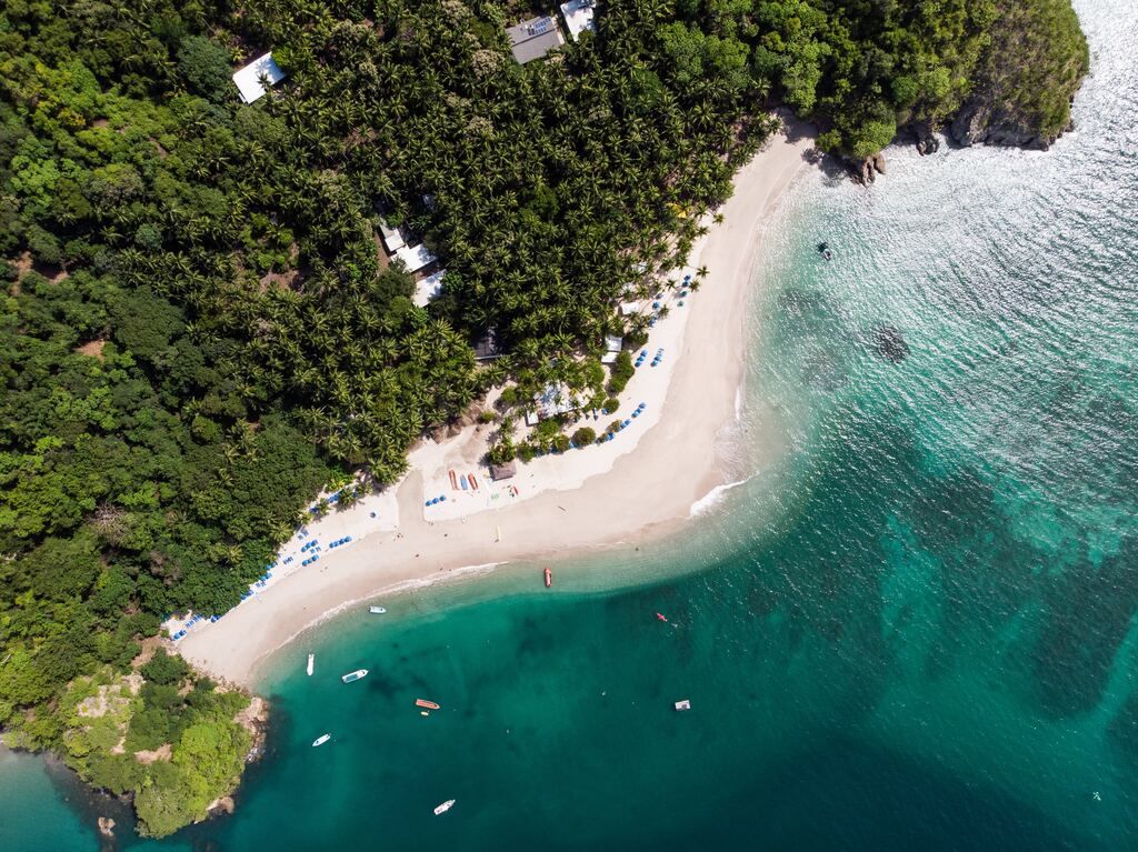 View of a white sand beach in Costa Rica, bordered by a dense palm forest and turquoise waters with small boats.