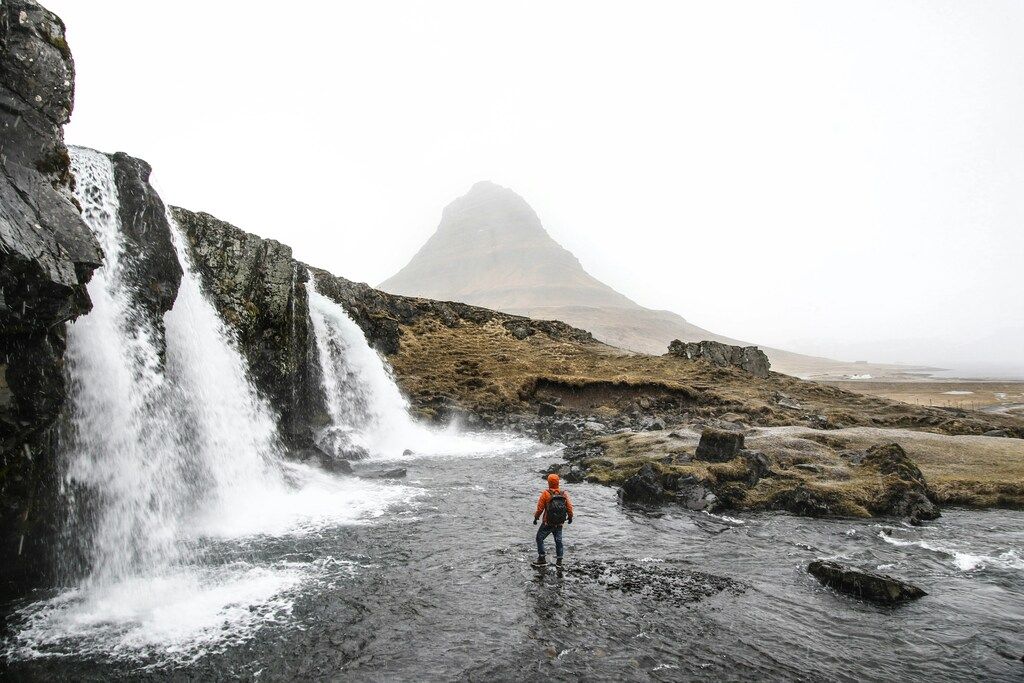 Man with backpack standing on river beside waterfall.