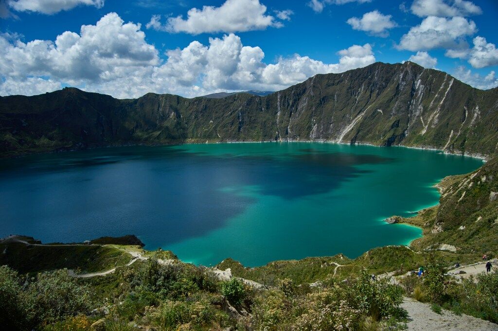 A wide view of the turquoise Quilotoa crater lake surrounded by steep volcanic walls under a bright blue sky.
