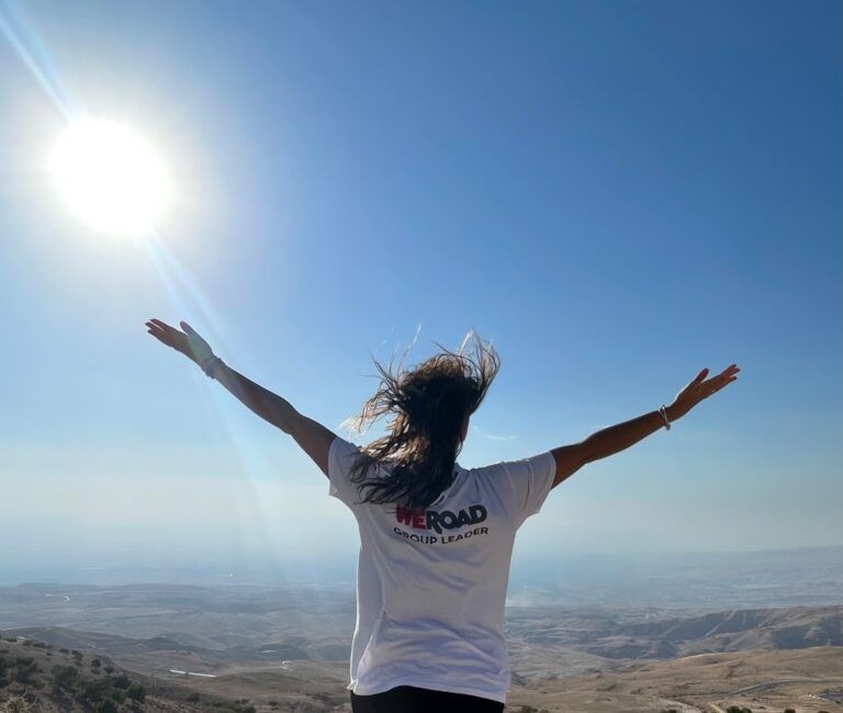 A WeRoad group leader stands with open arms facing a bright sun over a vast desert landscape in June.