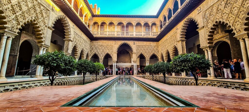 The stunning Courtyard of the Maidens in the Alcázar of Seville, featuring intricate Moorish arches and a long reflecting pool.