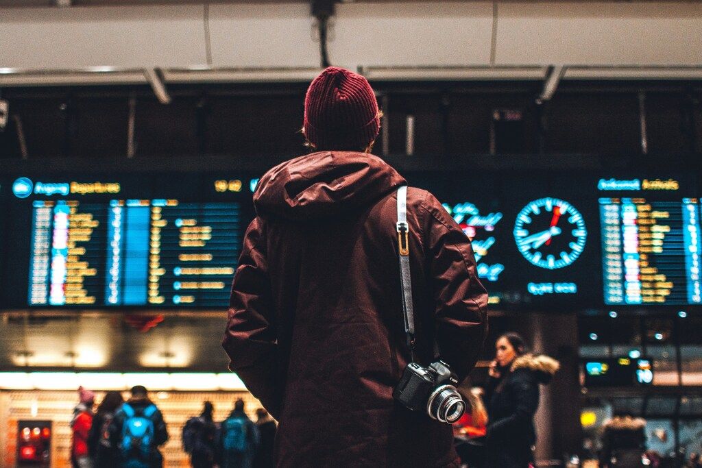 A person stands facing a flight information display board in an airport