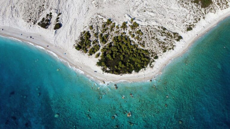 Drone shot of a white sandy beach with turquoise water and a small patch of green trees.
