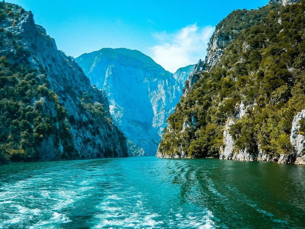 A boat's eye view traveling through a deep river canyon in Albania with steep, rocky cliffs covered in green vegetation.