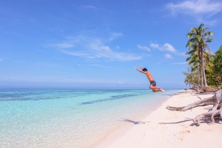 A person joyfully jumps into the clear turquoise water from a pristine white sand beach