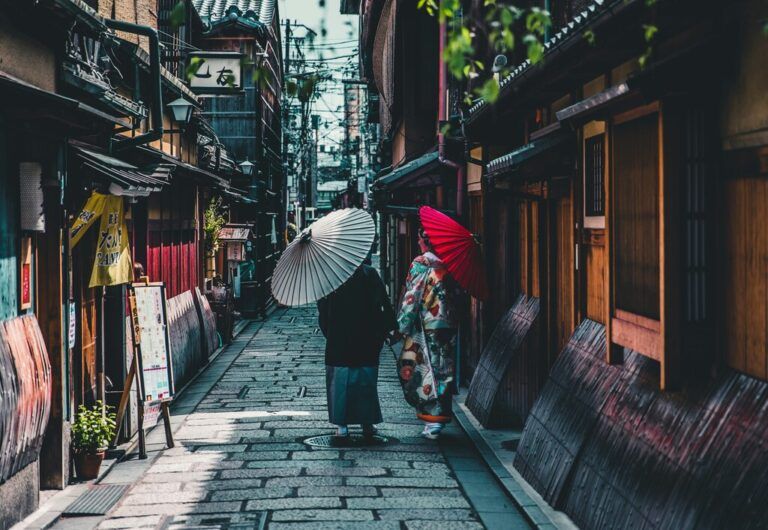 Two people dressed in traditional Japanese attire walk down a narrow street in Kyoto, holding decorative umbrellas.