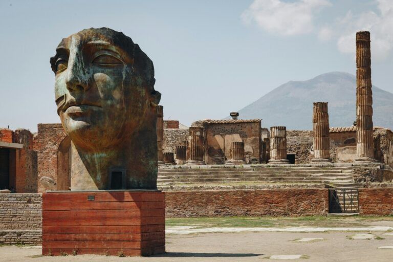 A large bronze statue of a face stands prominently in the ancient ruins of Pompeii