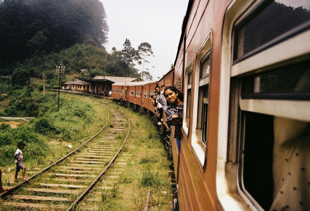 Passengers leaning out of the windows of a moving train as it curves through the lush green landscape of Sri Lanka.