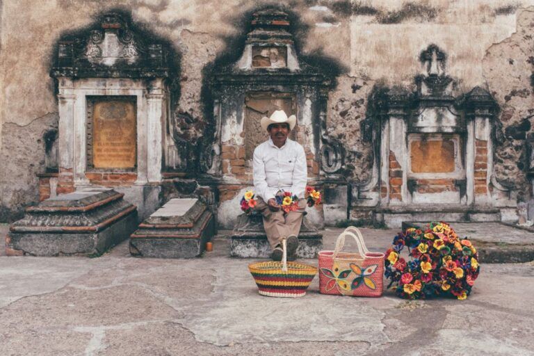 A man in traditional Maxica attire sits in front of ancient gravestones
