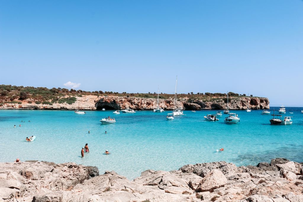 Crystal clear turquoise water at a sandy beach with several boats anchored and people swimming under a bright blue sky.