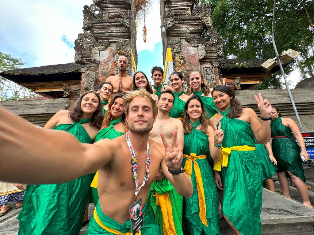 WeRoad travelers pose for a group selfie in Indonesia, wearing matching green and yellow sarongs in front of a Balinese temple gate.