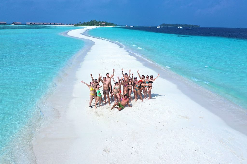 High-angle view of a group of WeRoad travelers posing on a narrow white sandbar surrounded by turquoise water.