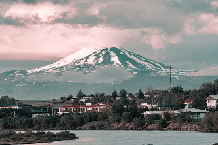 Snow-capped Hekla volcano towering over a small, low-rise village and a body of water in Iceland.