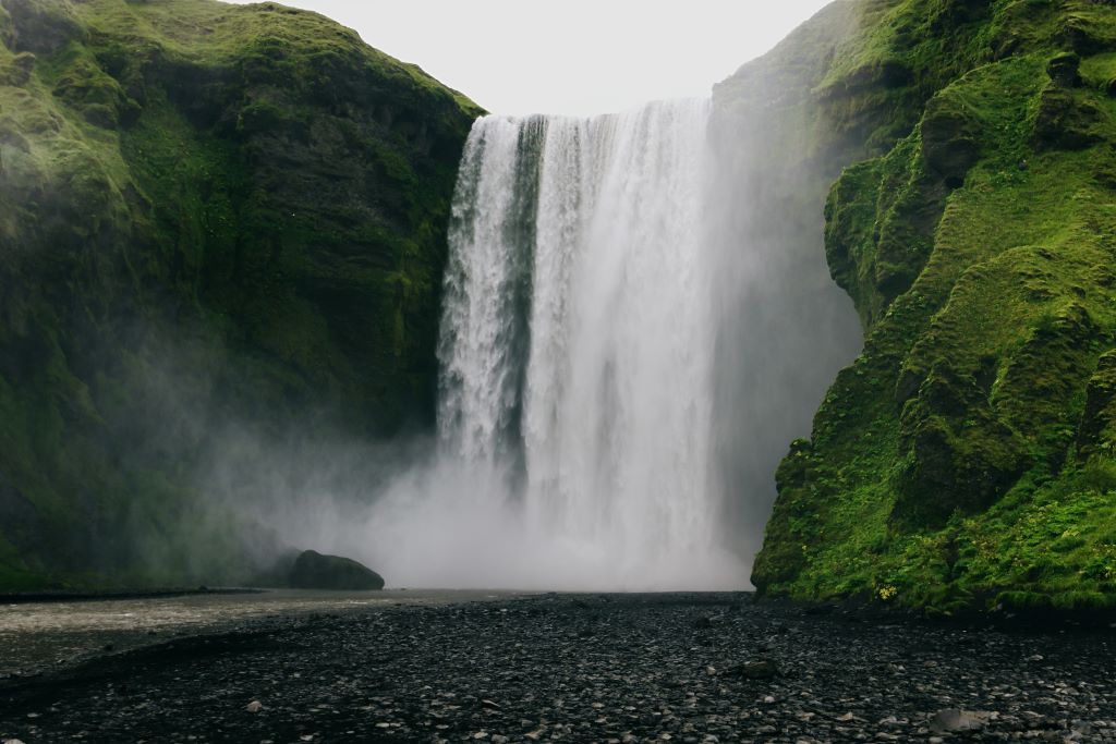 Wide view of the Skógafoss waterfall crashing over a high cliff onto a black pebble beach.