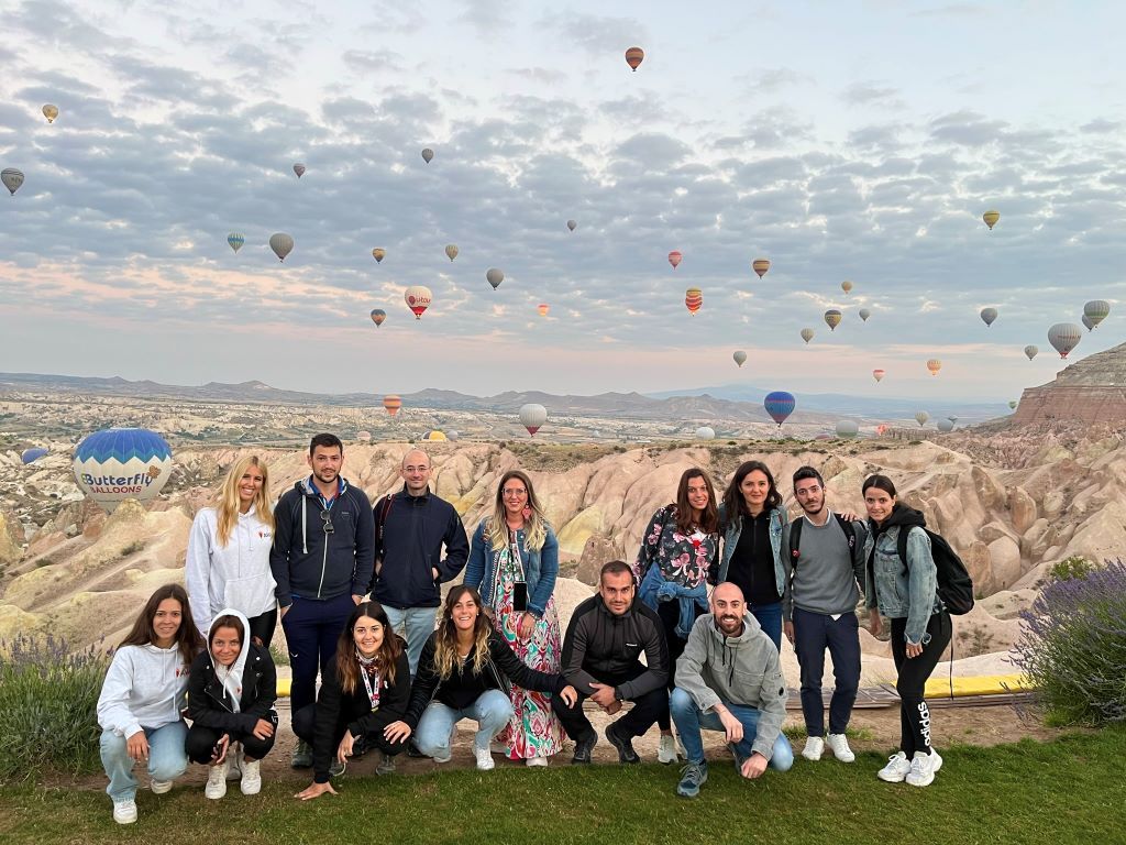 A group of WeRoad travelers posing on a hill in Cappadocia while dozens of hot air balloons fly in the sky at dawn.