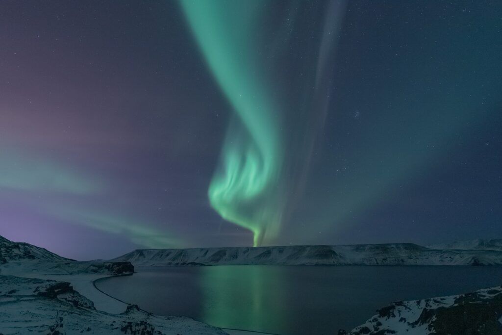 A vibrant green aurora borealis swirling in the starry night sky over a calm lake and snow-covered mountains.