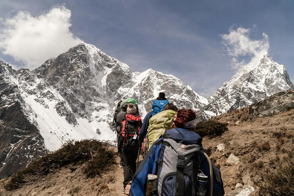 A group of hikers with large backpacks trekking along a mountain path toward towering, snow-capped Himalayan peaks.