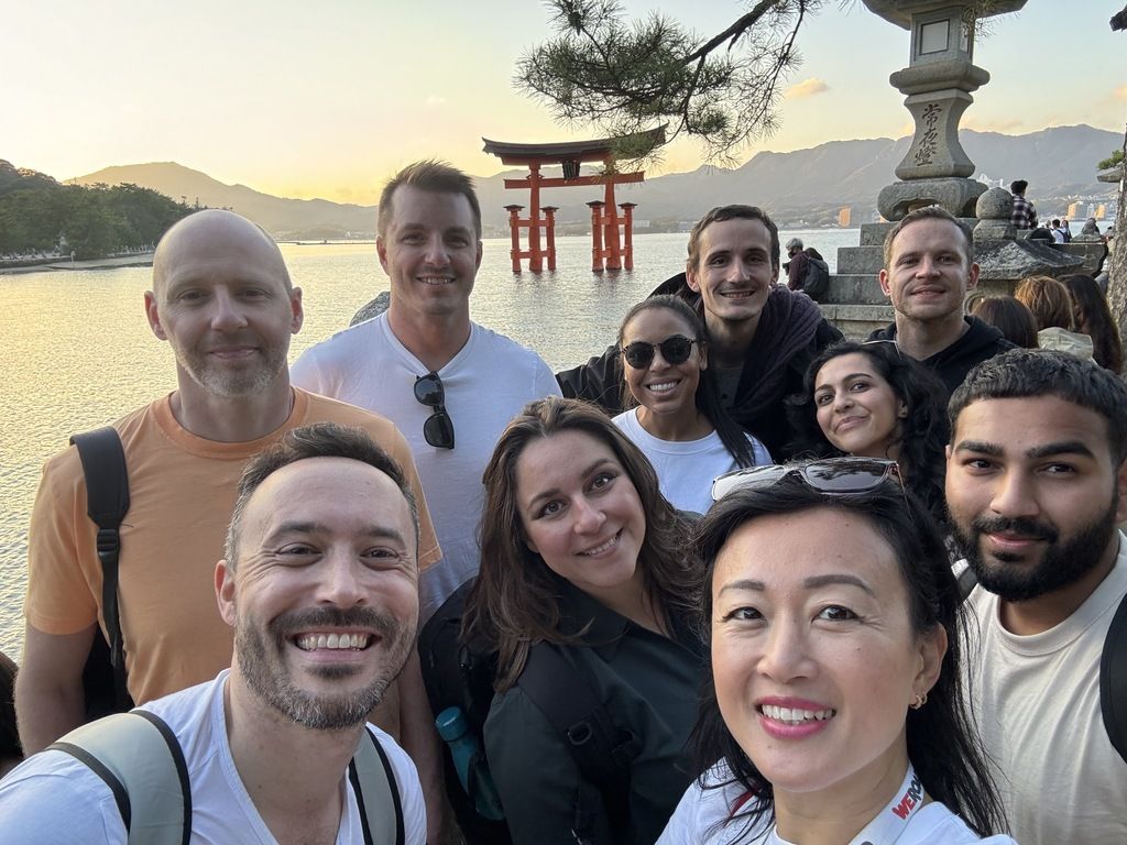 A group of WeRoad travelers smiles for a selfie with the famous floating Torii gate of Itsukushima Shrine in the background at sunset.