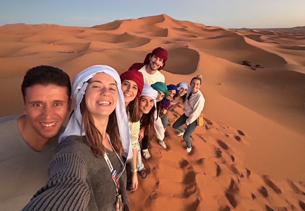 A cheerful group of WeRoad travelers taking a selfie on the orange sand dunes of the Sahara Desert in Morocco at sunset.