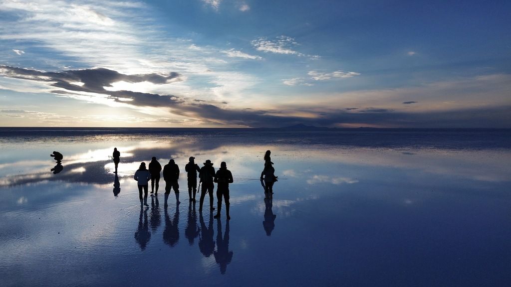  A group of people standing on the mirror-like surface of the salt flats during sunset.