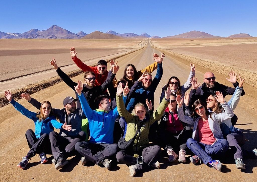 A group of WeRoad travelers sitting on a long, straight desert road in Chile, waving happily with mountains in the background.