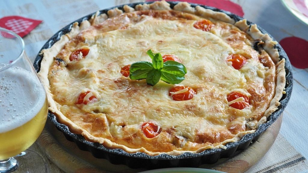 A golden Quiche Lorraine with cherry tomatoes and fresh basil, served with a glass of beer.