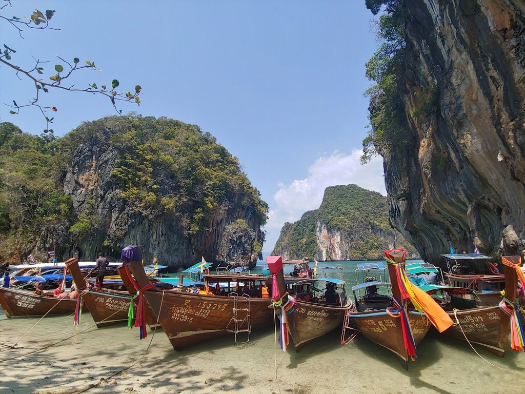 Traditional wooden longtail boats docked on a pristine beach in Thailand, with limestone cliffs in the background.