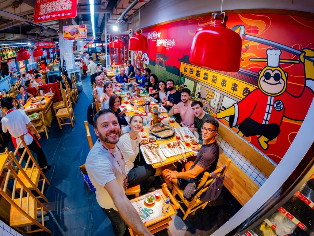 A group of WeRoad travelers smiling for a selfie while dining together at a lively, brightly lit Chinese restaurant.