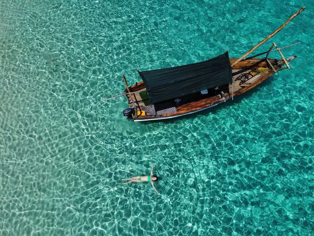 Traditional wooden dhow floating on clear turquoise waters, with a traveler relaxing in the sea below.