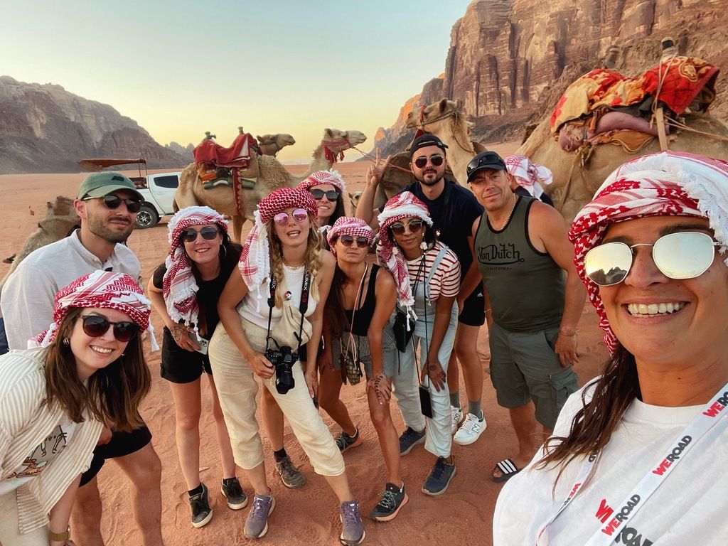 WeRoad travelers wearing keffiyehs posing with camels in Wadi Rum desert, Jordan.
