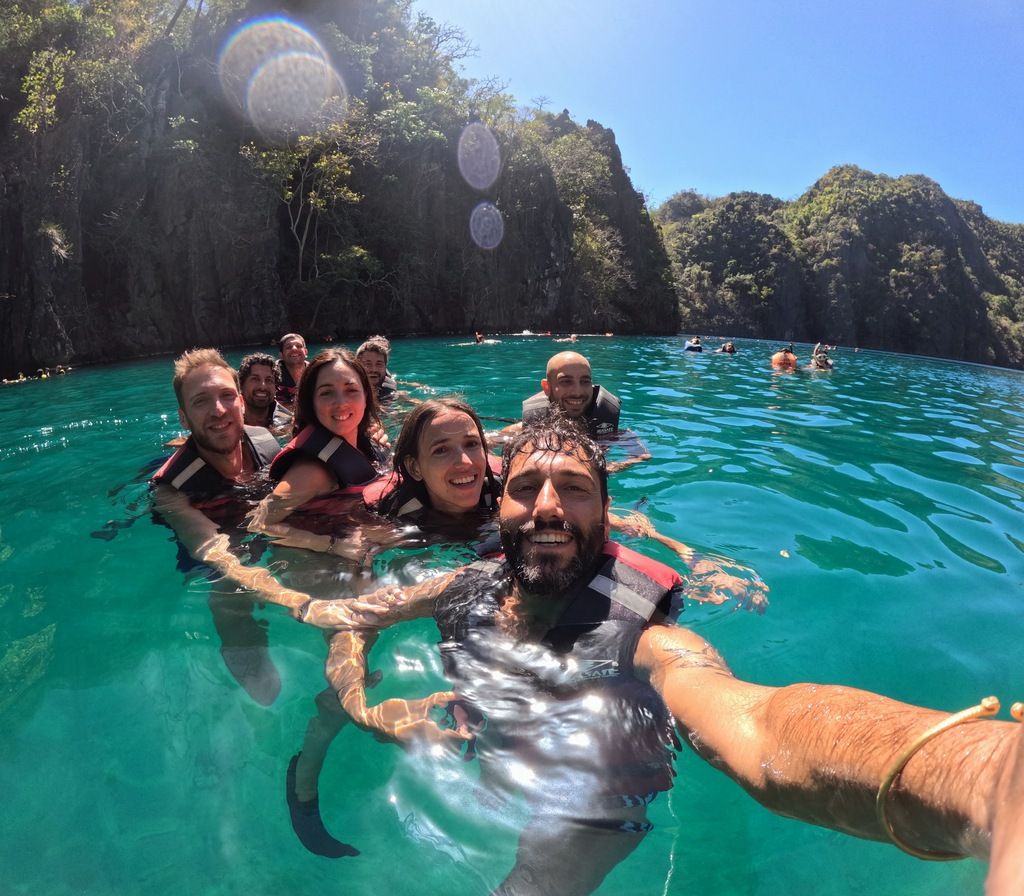 WeRoad travelers group swimming and smiling in crystal clear turquoise water, surrounded by towering limestone cliffs in the Philippines.
