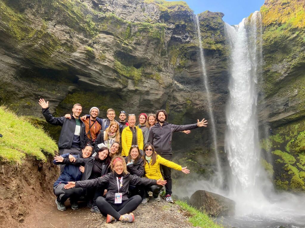 Smiling group of WeRoad travelers posing in front of a tall waterfall in Iceland.
