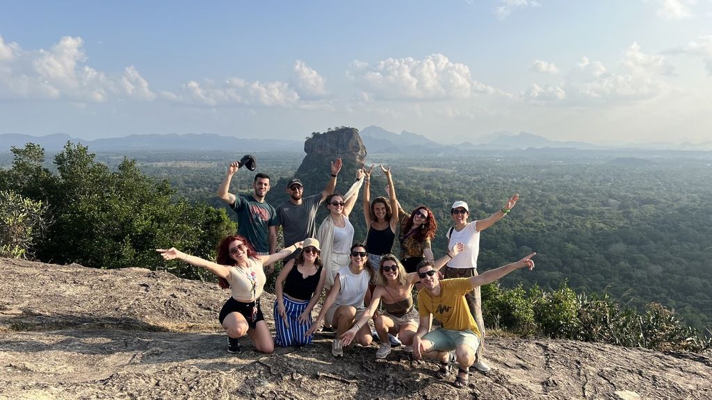 A group of happy WeRoad travelers posing on a rocky peak with the ancient Sigiriya rock fortress in the background.