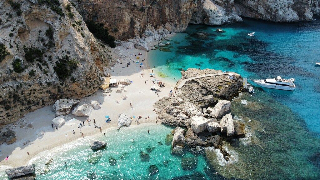View of a secluded white sand beach with turquoise waters and white yachts anchored near rocky cliffs.