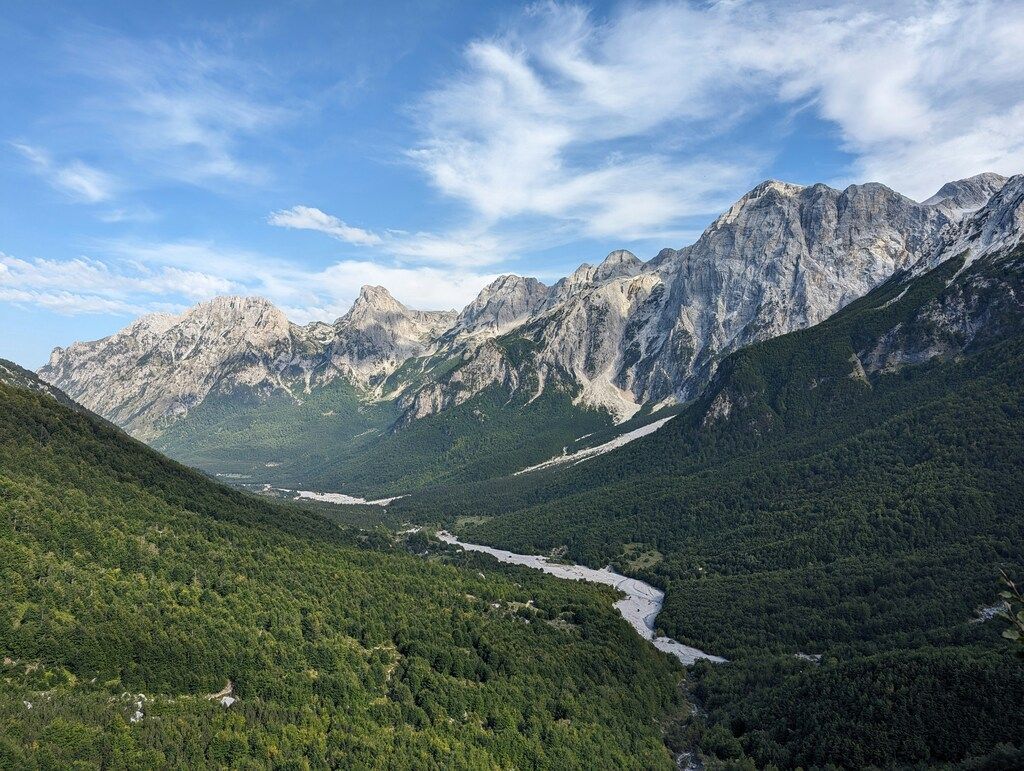 A breathtaking view of the Valbona Valley in Albania, featuring a winding riverbed at the base of towering, lush green mountains under a bright blue sky.