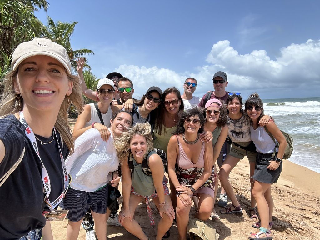A large group of smiling WeRoad travelers taking a selfie on a sunny beach in Albania with the sea in the background.
