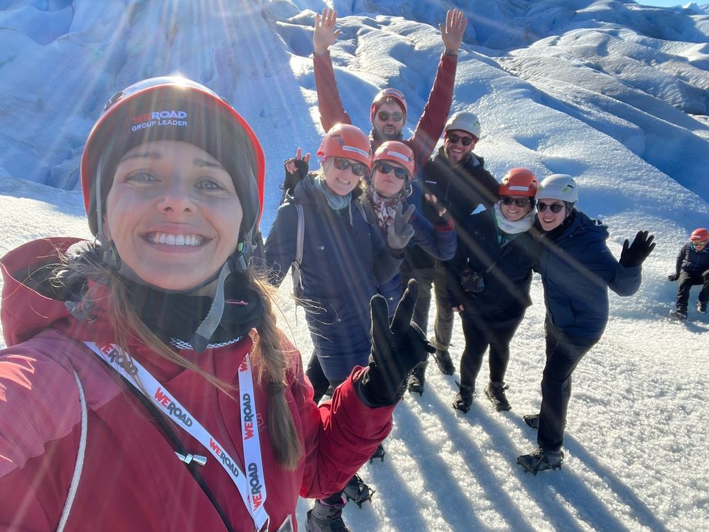 A group of smiling WeRoad travelers wearing helmets and crampons taking a selfie on a bright, sunny glacier in Patagonia.