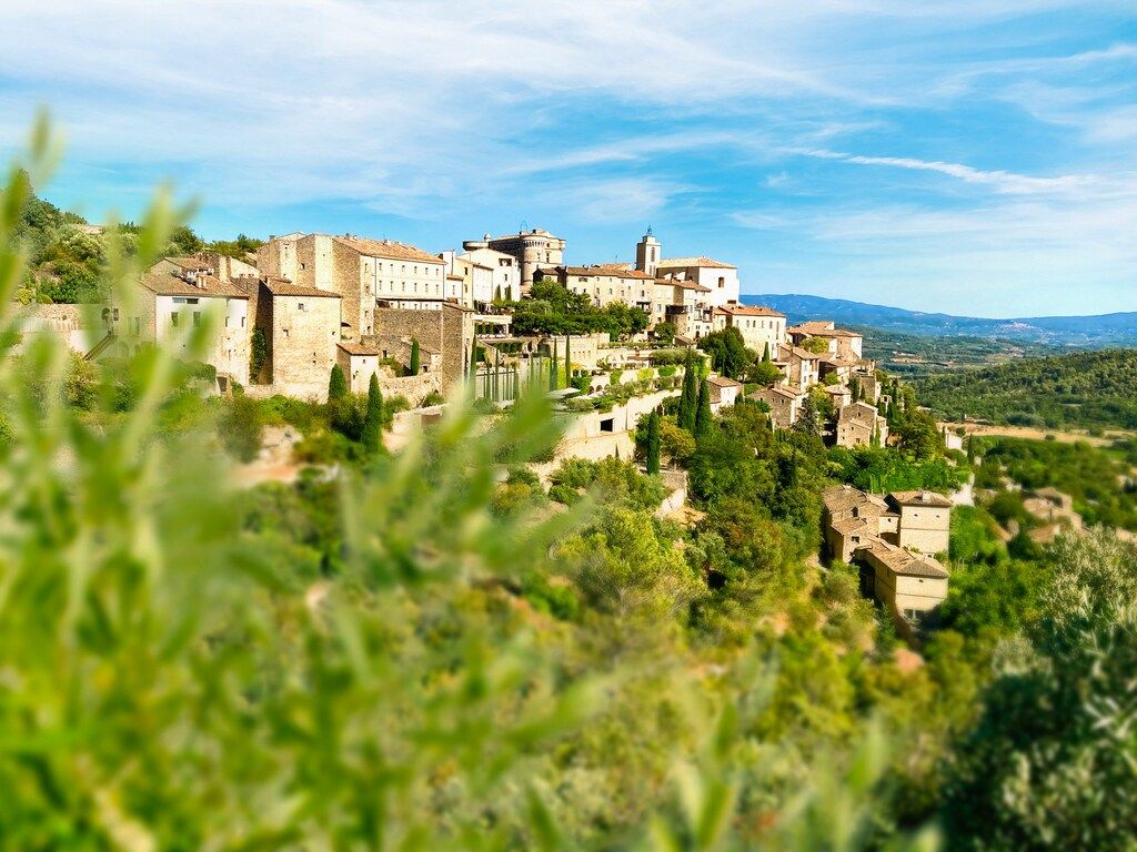 A picturesque hilltop village with stone buildings and red roofs, surrounded by lush green trees and distant mountains under a blue sky with some clouds.