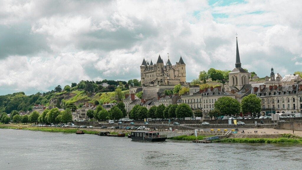 A large, historic château with multiple turrets and spires on a hill overlooking a wide river, with a town built along the riverbank.