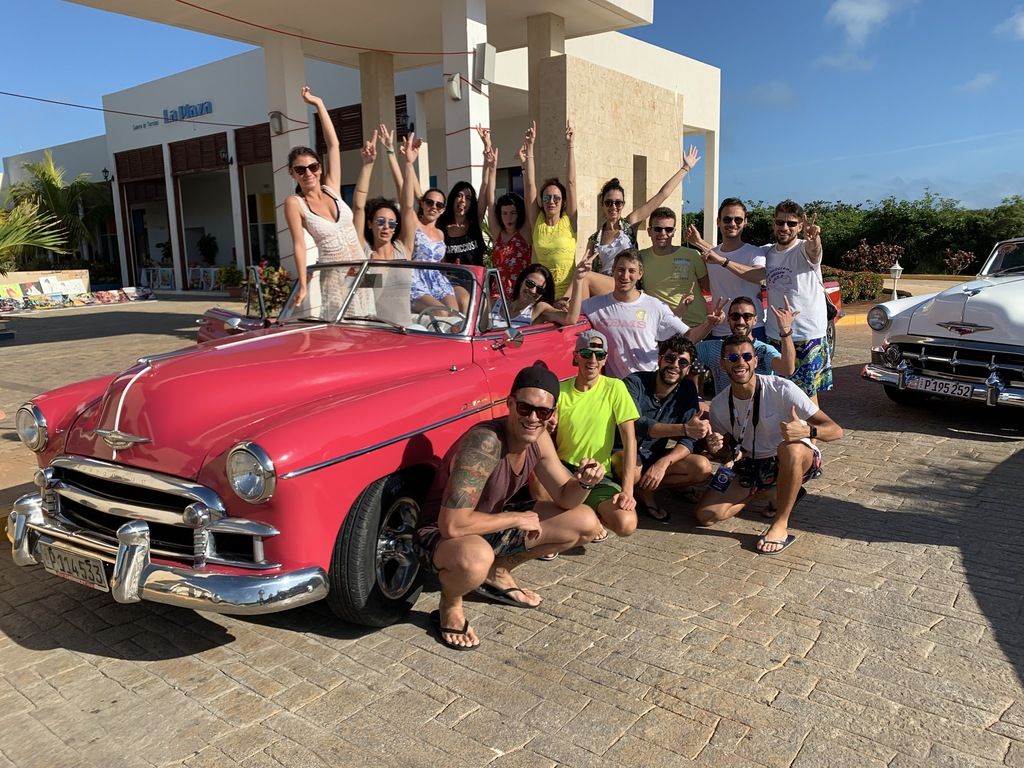 A group of happy WeRoad travelers posing with a classic pink vintage convertible and a white classic car in a sunny outdoor setting in Cuba.