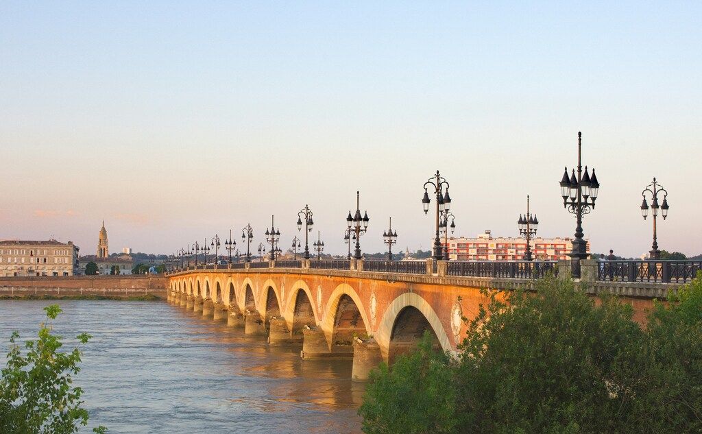 The Pont de Pierre bridge in Bordeaux, France, with its many arches and lampposts spanning a river at sunset, with city buildings on the far bank.