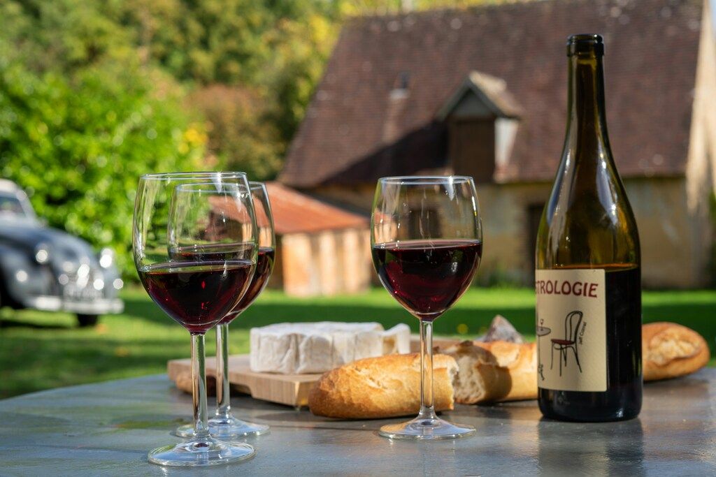 Two glasses of red wine, a bottle of red wine, bread, and cheese on an outdoor table with a historic building and car in the background.