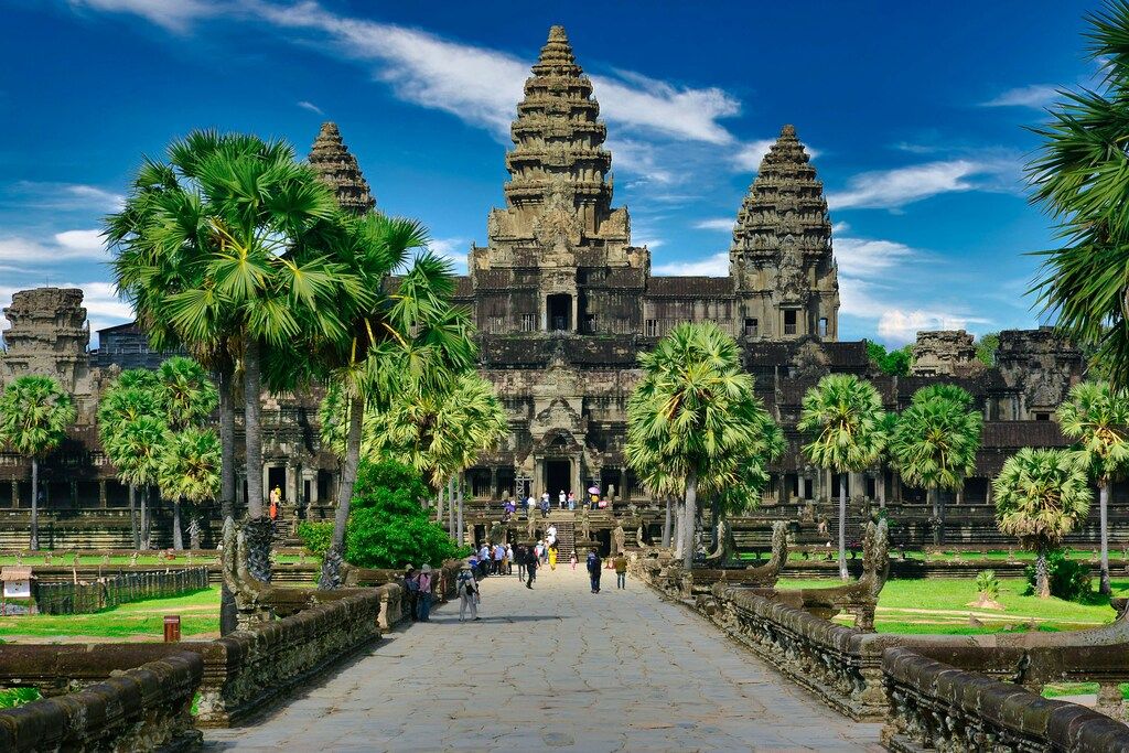 The ancient stone temple complex of Angkor Wat in Cambodia, surrounded by green trees and a wide pathway leading to its grand entrance under a blue sky with white clouds.