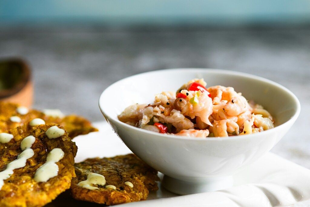 A bowl of ceviche with shrimp or seafood, accompanied by fried plantain slices (tostones) with a creamy sauce.