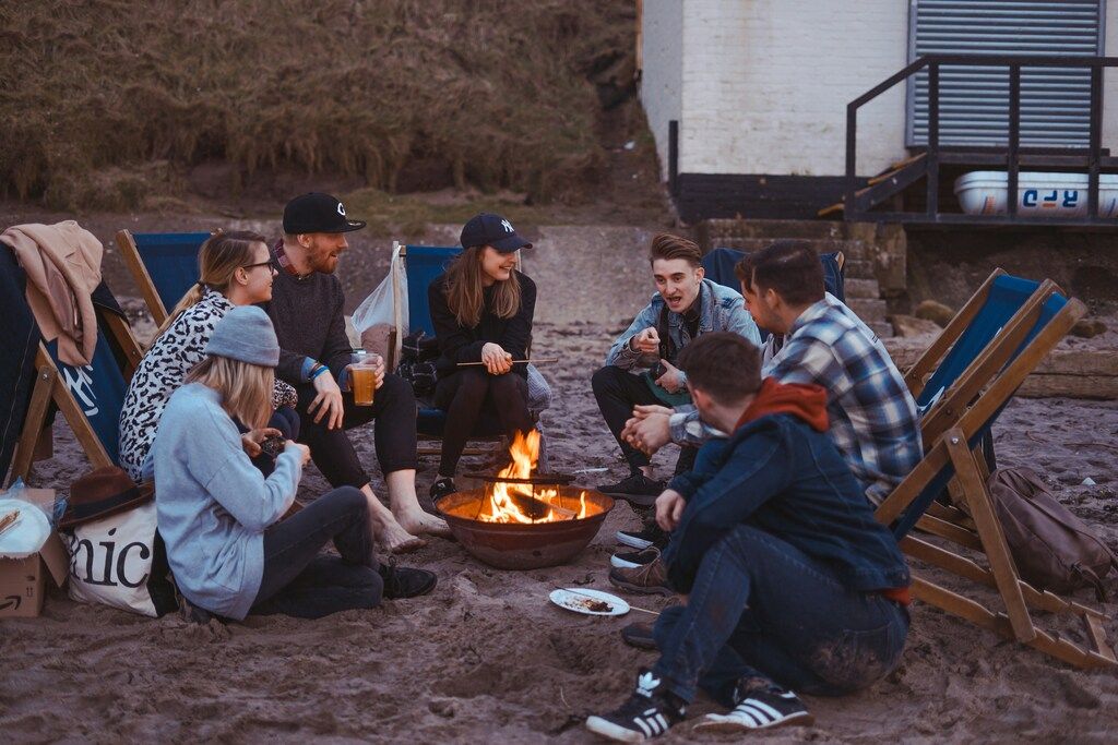 A group of young adults sits around a bonfire on a sandy beach at dusk. 