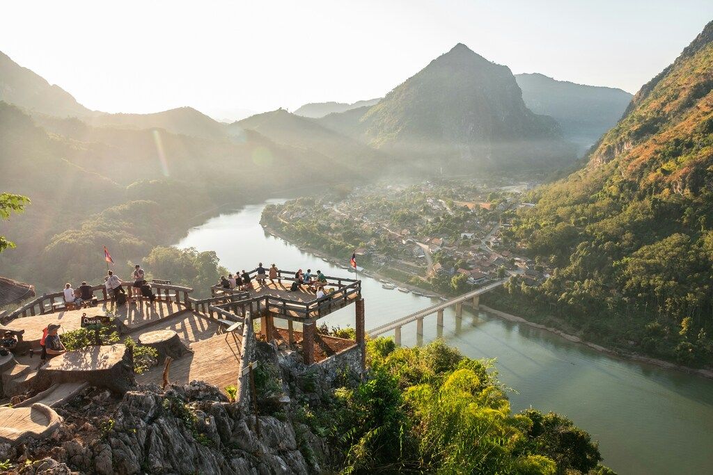A viewpoint with people overlooking a river winding through a valley, a town with a bridge, and mountains in the distance, bathed in sunlight.