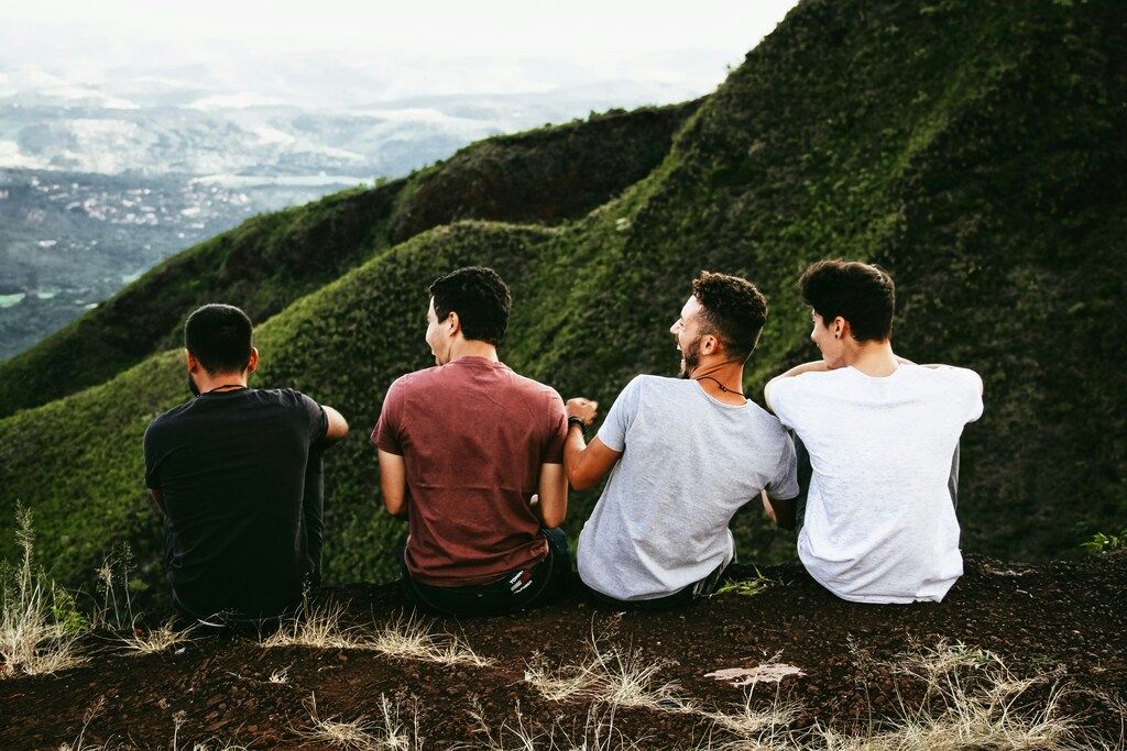 Four young men are sitting with their backs to the camera, looking out over a lush, green, mountainous landscape. 